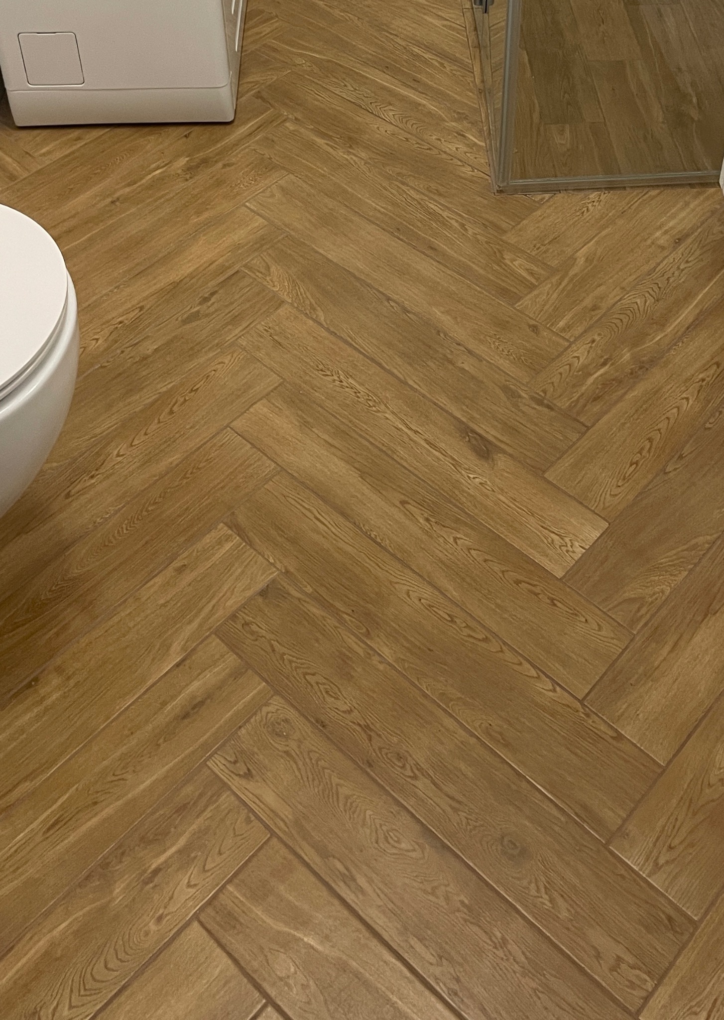 Ułożona w jodełkę podłoga z płytek imitujących drewno w łazience, widoczny fragment toalety i kabiny prysznicowej. Ciepłe oświetlenie podkreśla strukturę drewna.