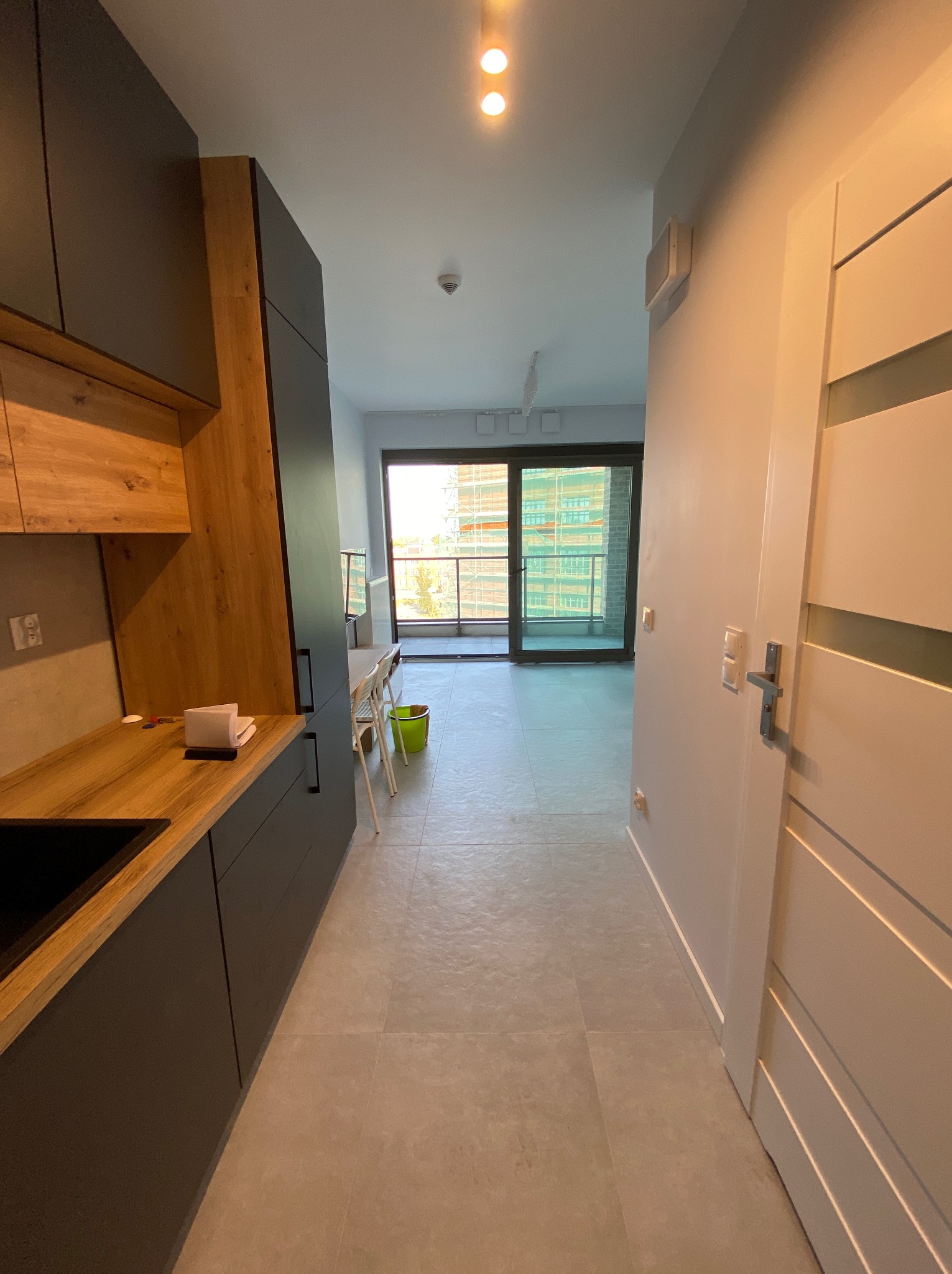 Nowoczesna kuchnia z drewnianymi akcentami i szarymi szafkami, widok na jasny salon z balkonem. Minimalistyczny design, świeże wykończenie, płytki podłogowe w odcieniach szarości.