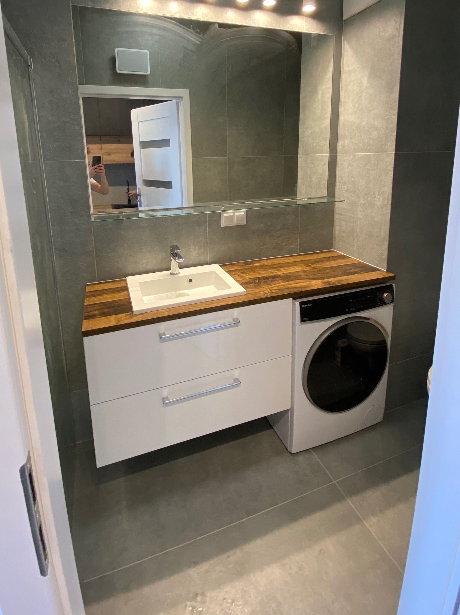 Nowoczesna łazienka z szarymi płytkami, drewnianym blatem, białą umywalką i pralką. W lustrze odbija się fragment innego pomieszczenia.