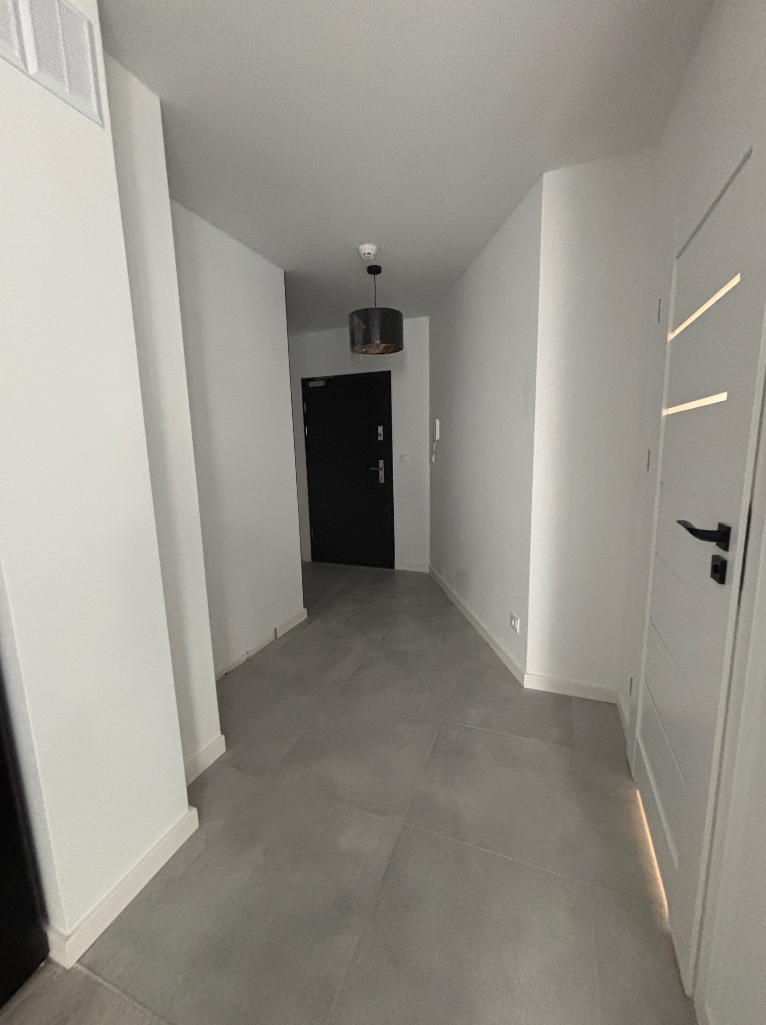 Nowoczesny korytarz z szarymi płytkami podłogowymi, białymi ścianami i czarnymi drzwiami wejściowymi. Minimalistyczna lampa sufitowa i białe drzwi wewnętrzne z czarną klamką.