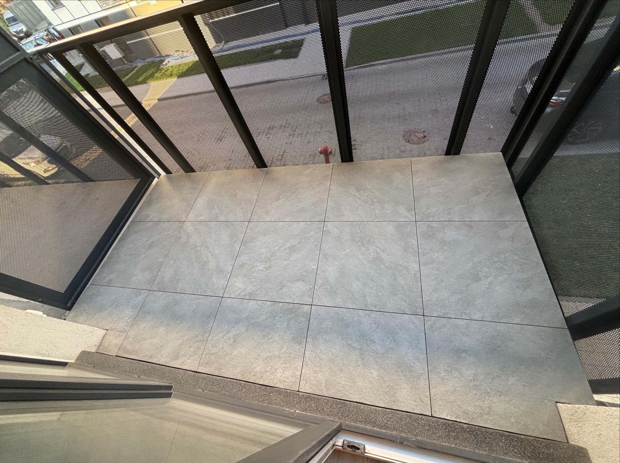 Wykończony balkon z szarymi płytkami podłogowymi i metalową balustradą z siatki, widok z góry, równomierne oświetlenie.