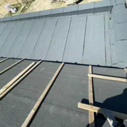 Częściowo pokryty papą dach z drewnianymi łatami, widok z góry, cień osoby w prawym dolnym rogu.