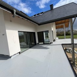 D-Dach Dawid Leszczyński - Nowoczesny dom z dachem pokrytym szarą dachówką i obróbkami blacharskimi. Widoczne okna dachowe i orynnowanie. Wykończony taras z szarą izolacją.