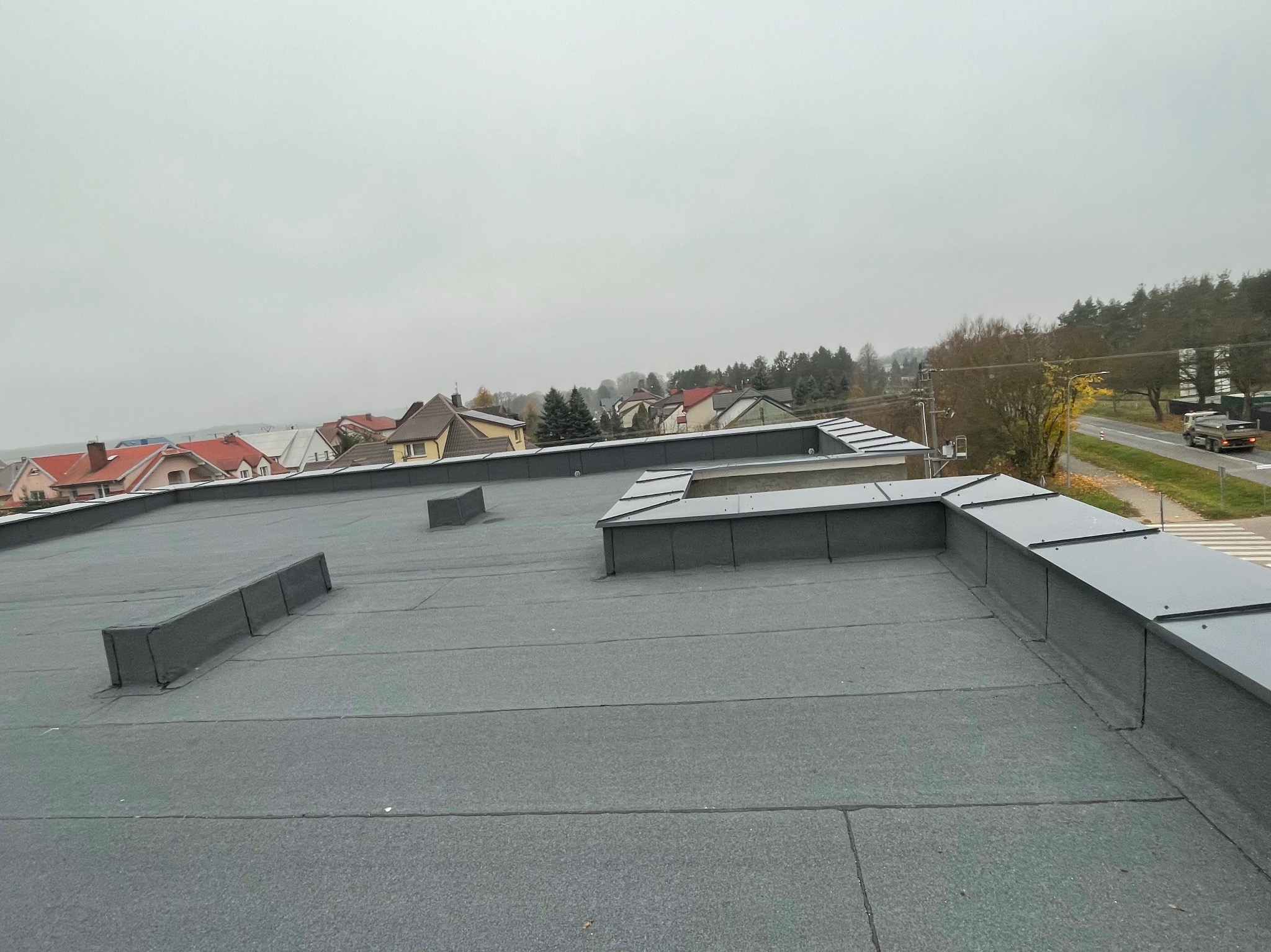 Płaski dach pokryty papą termozgrzewalną, widoczne kominki wentylacyjne i obróbki blacharskie na krawędzi dachu, w tle zabudowa jednorodzinna i pochmurne niebo.