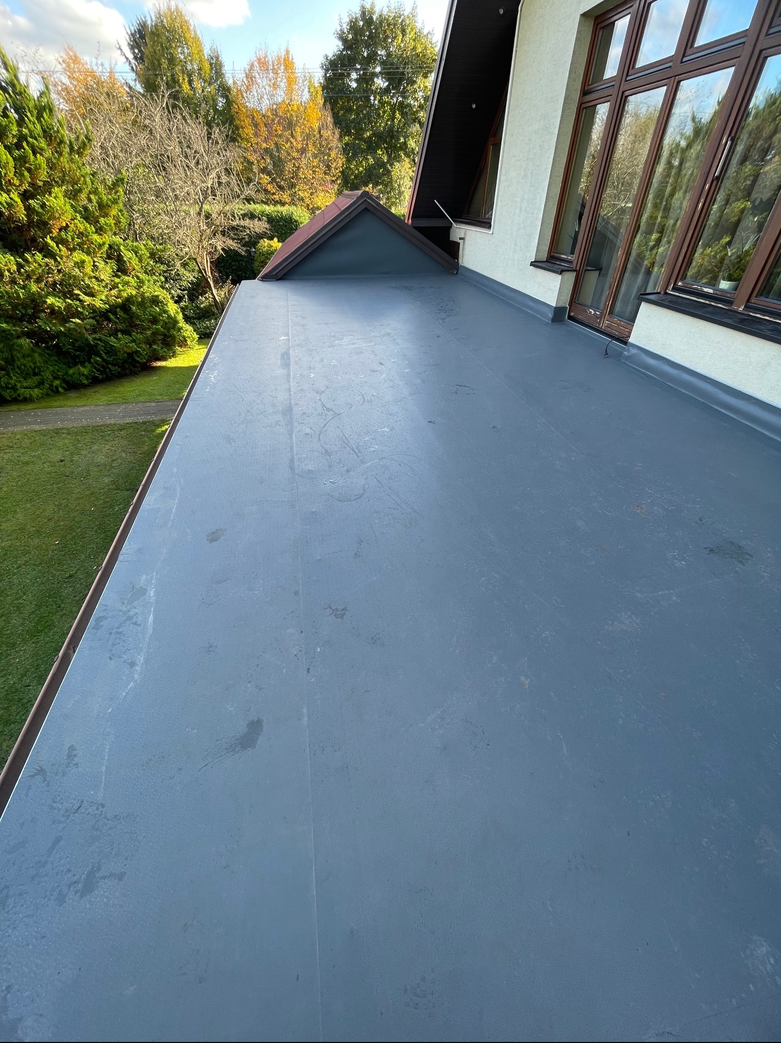 Widok z góry na płaski, szary dach pokryty papą termozgrzewalną, z widocznymi śladami po wodzie i lekkimi zabrudzeniami, w tle fragment budynku z oknami i zielenią.