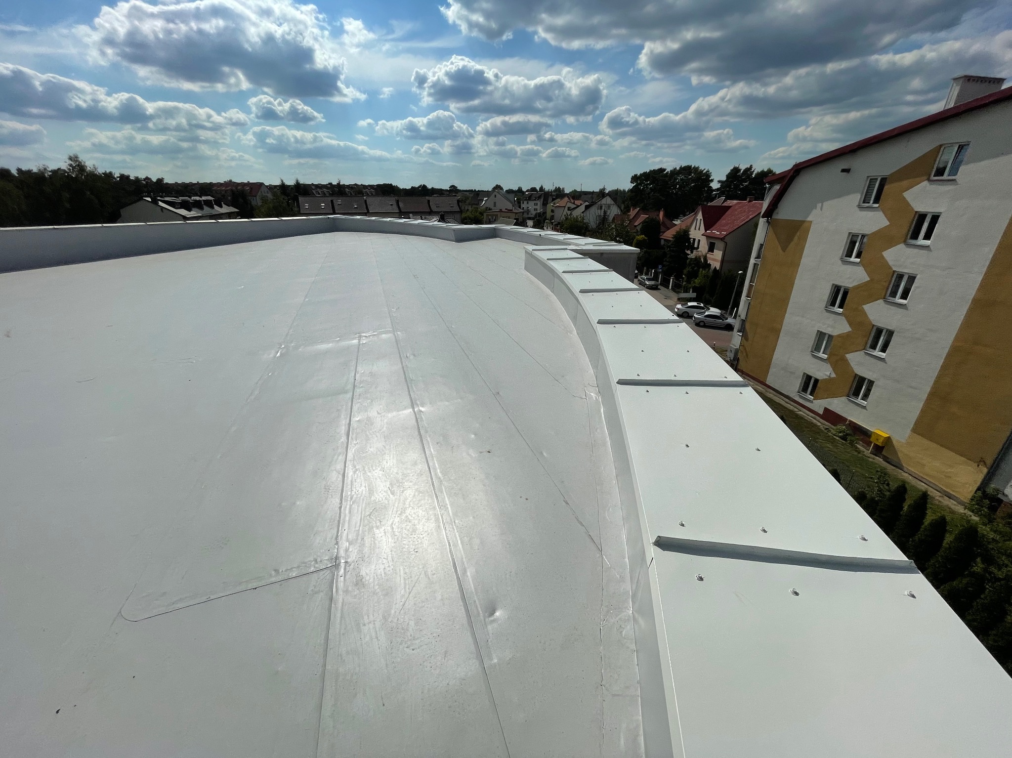 Biała, płaska powierzchnia dachu z widocznymi łączeniami i obróbkami blacharskimi, na tle zabudowy miejskiej i błękitnego nieba z chmurami.