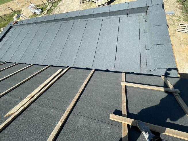 Częściowo pokryty papą dach z drewnianymi łatami, widok z góry, cień osoby w prawym dolnym rogu.