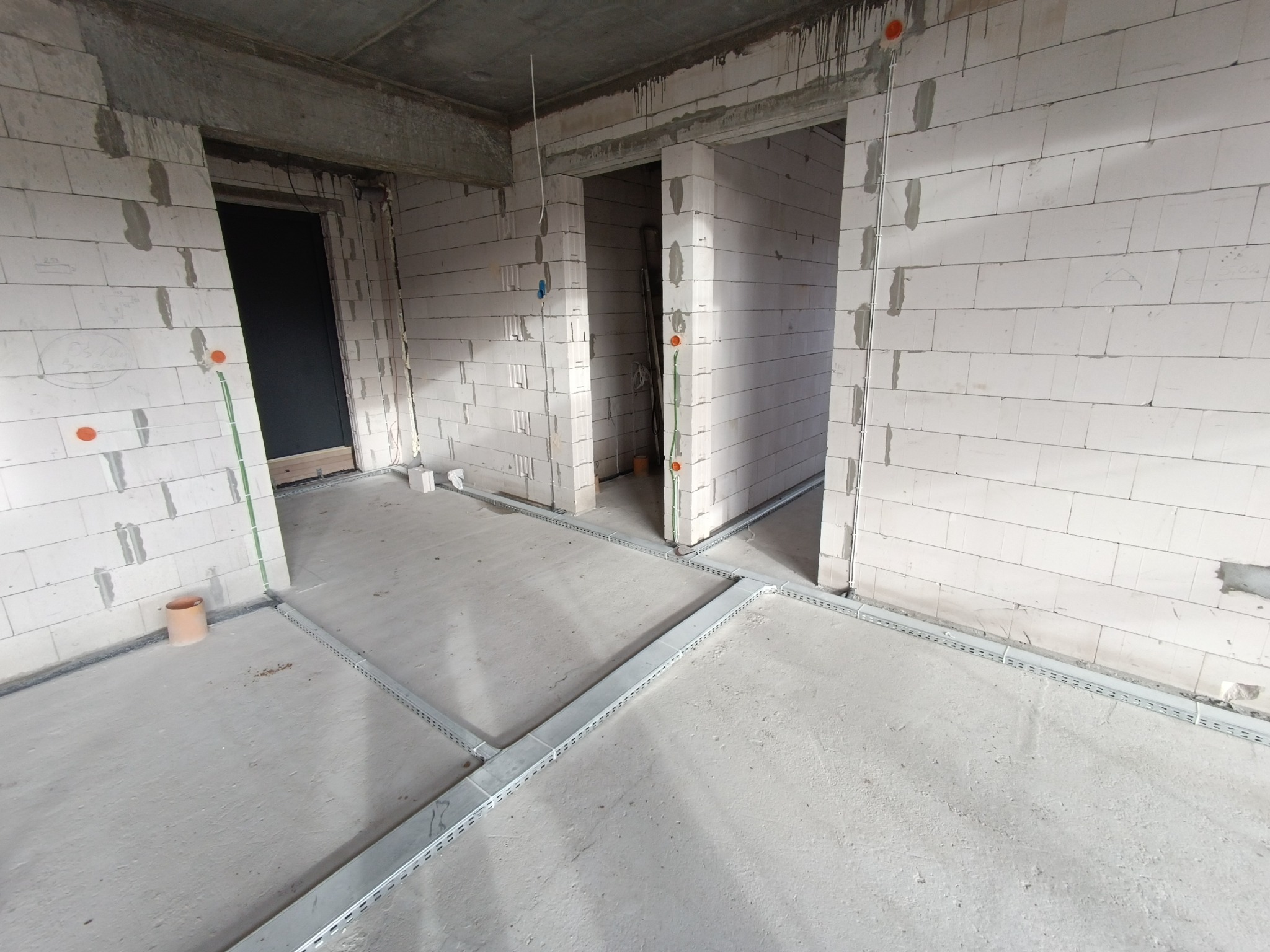 Surowy stan wnętrza z bloków, widoczne koryta kablowe na podłodze i pionowe peszle z puszkami elektrycznymi na ścianach.