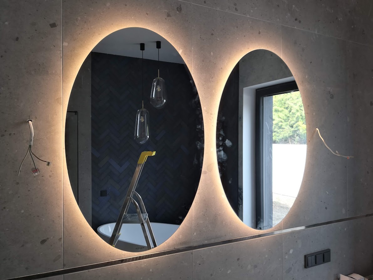 Dwa owalne lustra z podświetleniem LED na szarej, kamiennej ścianie w łazience, widoczne wystające kable i drabina w odbiciu, sugerujące prace instalacyjne.