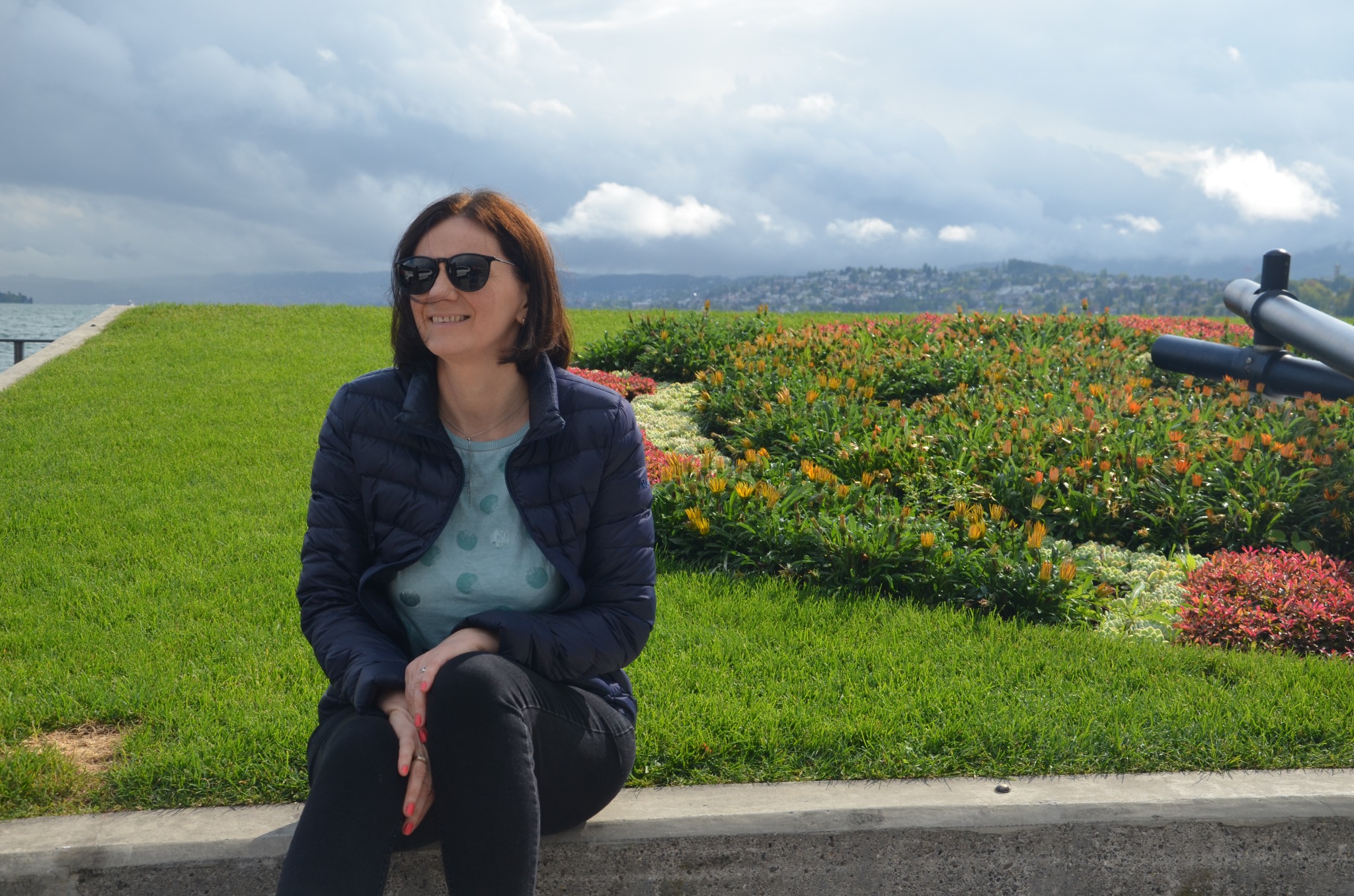 Uśmiechnięta kobieta w okularach przeciwsłonecznych siedzi na tle zieleni i kwiatów, z widokiem na jezioro i miasto w oddali, w słoneczny dzień.