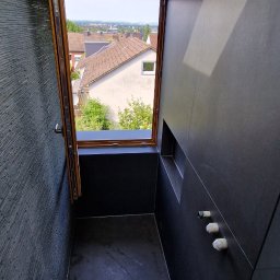 TPG BŁAŻEJ WOJDAN - Nowoczesna łazienka z czarnymi płytkami, oknem i odpływem liniowym. Widok na zabudowę miejską przez otwarte okno. Minimalistyczny design z elementami kamienia.