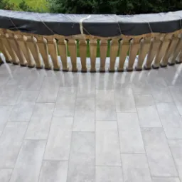 Jasnoszare, prostokątne płytki na balkonie z kamienną balustradą, zabezpieczoną czarną folią. Widok z góry na nową, błyszczącą posadzkę.