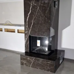 Nowoczesny kominek obłożony ciemnym marmurem z białymi żyłami, widoczny na tle surowego wnętrza z szarymi płytkami podłogowymi i fragmentem instalacji na ścianie.