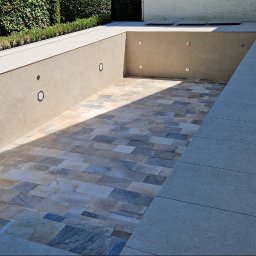 TPG BŁAŻEJ WOJDAN - Wykończony basen z mozaiką w odcieniach beżu i szarości, oświetleniem punktowym w ścianach, otoczony jasnymi płytami i zielenią. Widoczny odpływ liniowy.