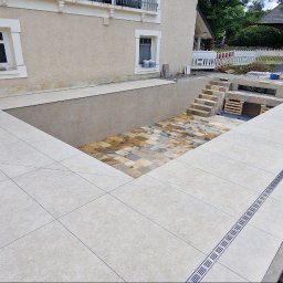 TPG BŁAŻEJ WOJDAN - Wykończony taras z beżowych płytek z odpływem liniowym. W tle zejście do ogrodu z kamienną posadzką i schodami. Prace wykończeniowe w toku.