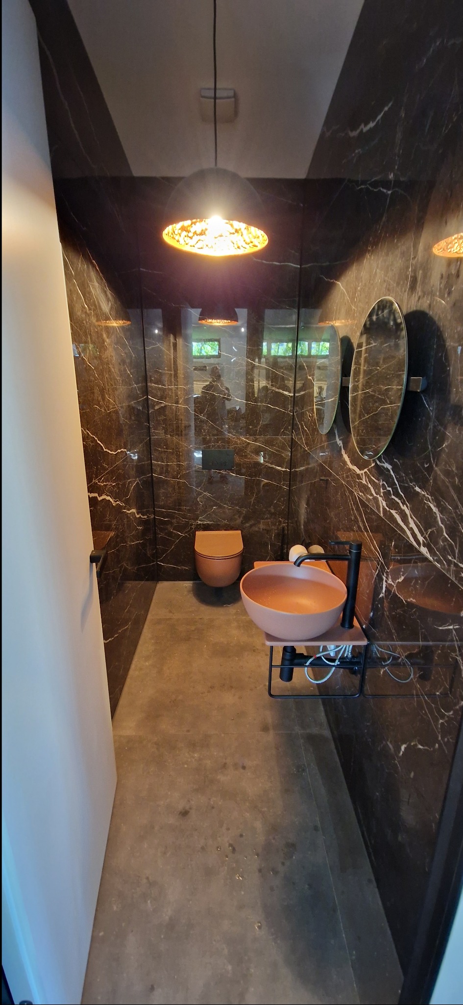 Nowoczesna łazienka z marmurowymi ścianami, okrągłymi lustrami i designerską umywalką w odcieniach terakoty. Lampa sufitowa z ażurowym kloszem. Widok na toaletę.