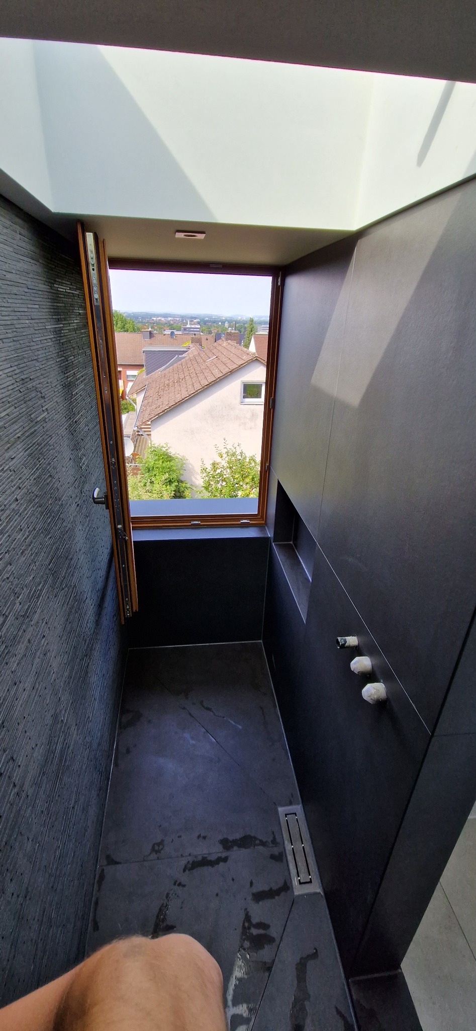 Nowoczesna łazienka z czarnymi płytkami, oknem i odpływem liniowym. Widok na zabudowę miejską przez otwarte okno. Minimalistyczny design z elementami kamienia.