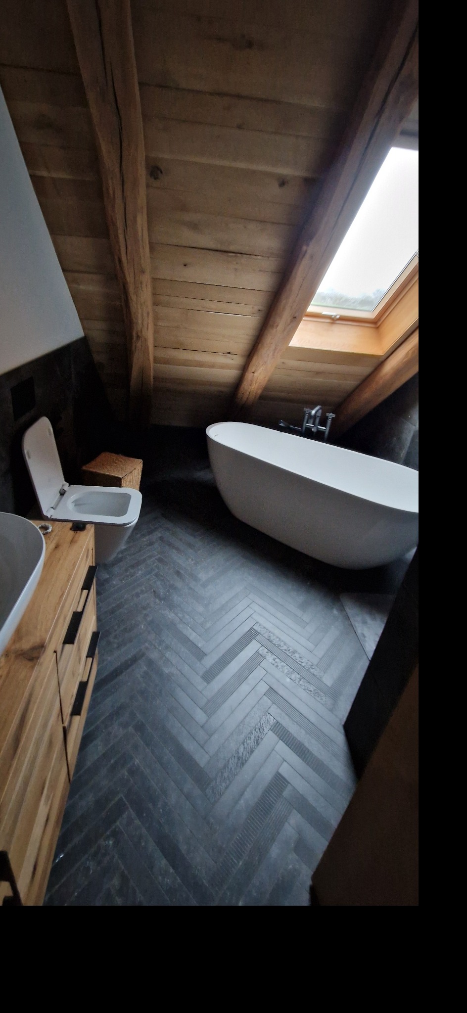Stylowa łazienka na poddaszu z drewnianym sufitem, oknem dachowym i czarną podłogą w jodełkę. Wolnostojąca wanna i umywalka z drewnianą szafką.