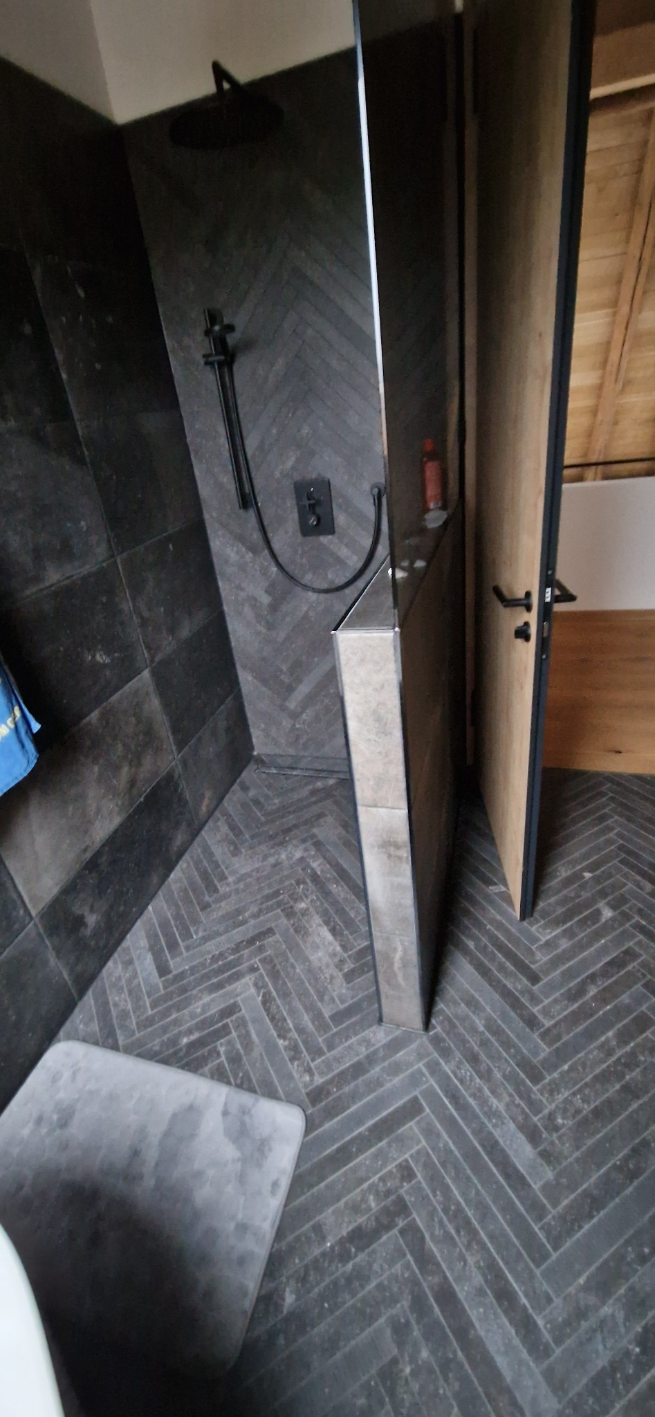 Nowoczesna łazienka z ciemnymi płytkami ułożonymi w jodełkę na ścianach i podłodze. Widoczna kabina prysznicowa i fragment drewnianych drzwi.