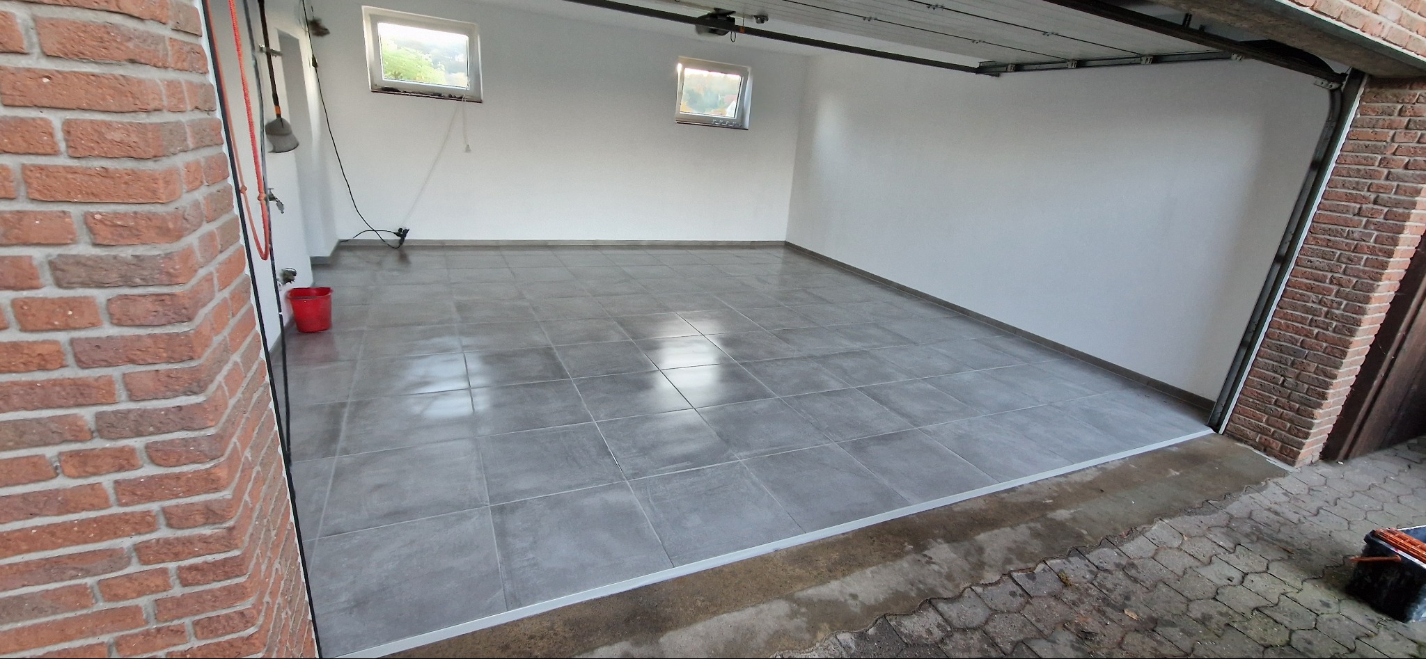 Wykończona podłoga z szarych, kwadratowych płytek w garażu. Widoczne białe ściany i ceglana kolumna przy otwartych drzwiach garażowych. Minimalistyczny wystrój.