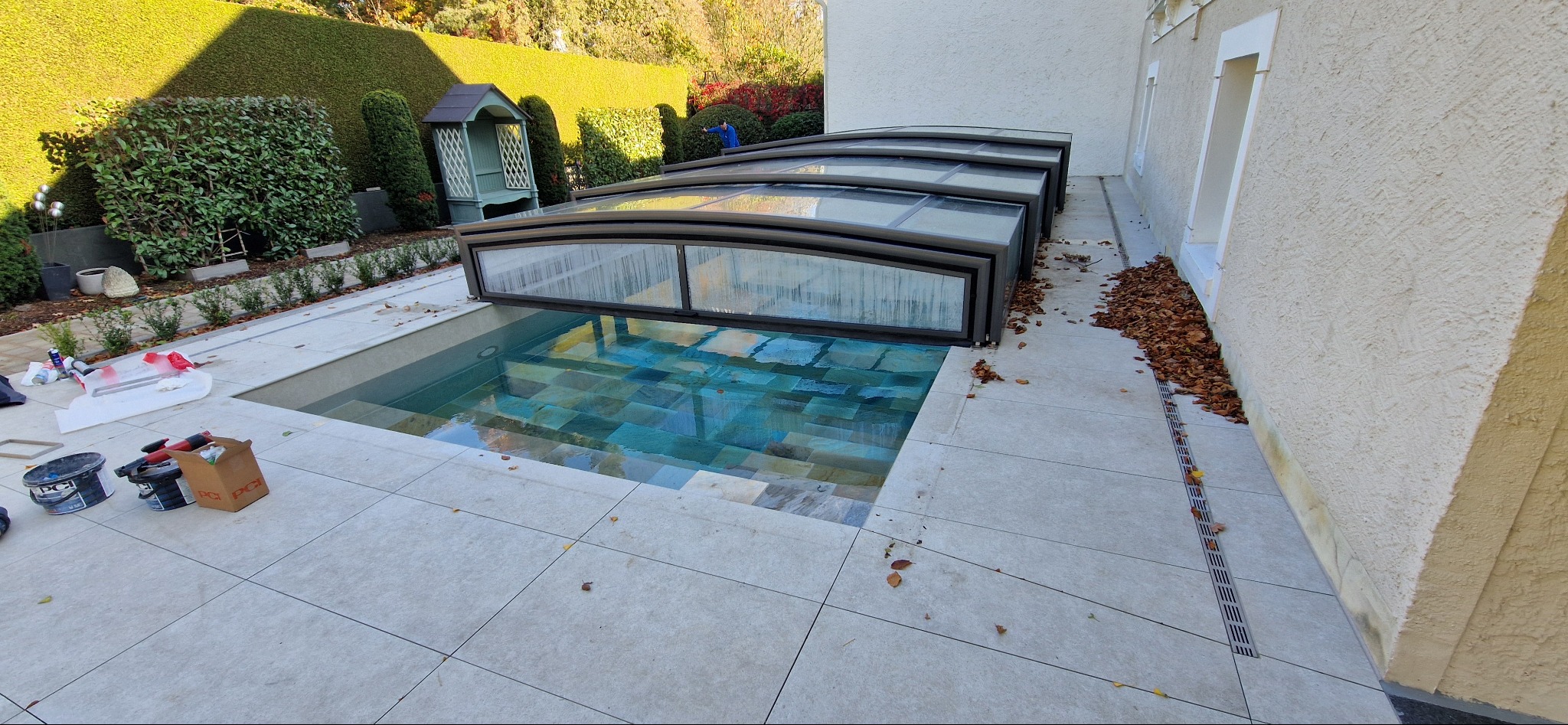 Zadaszony basen z mozaiką i jasnymi płytkami na zewnątrz. Widoczne narzędzia i materiały budowlane. Tło: zadbany ogród i elewacja budynku.