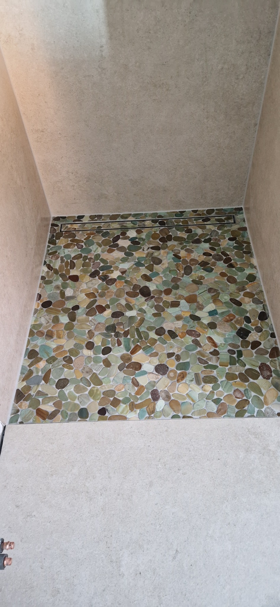 Wykończona kabina prysznicowa z odpływem liniowym i podłogą wyłożoną mozaiką z naturalnych kamieni w odcieniach beżu, brązu i zieleni, z jasnymi fugami.