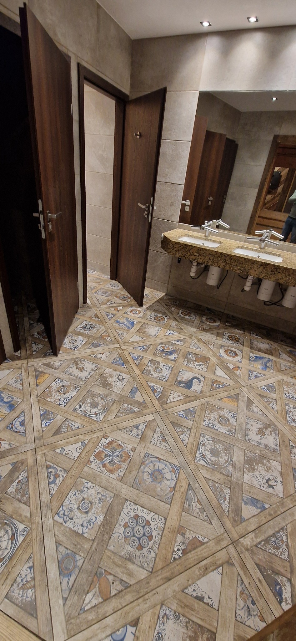 Wykończona łazienka z wzorzystą podłogą w stylu rustykalnym, drewnianymi drzwiami i umywalką z kamiennym blatem. Widoczne detale wykończenia i armatura.