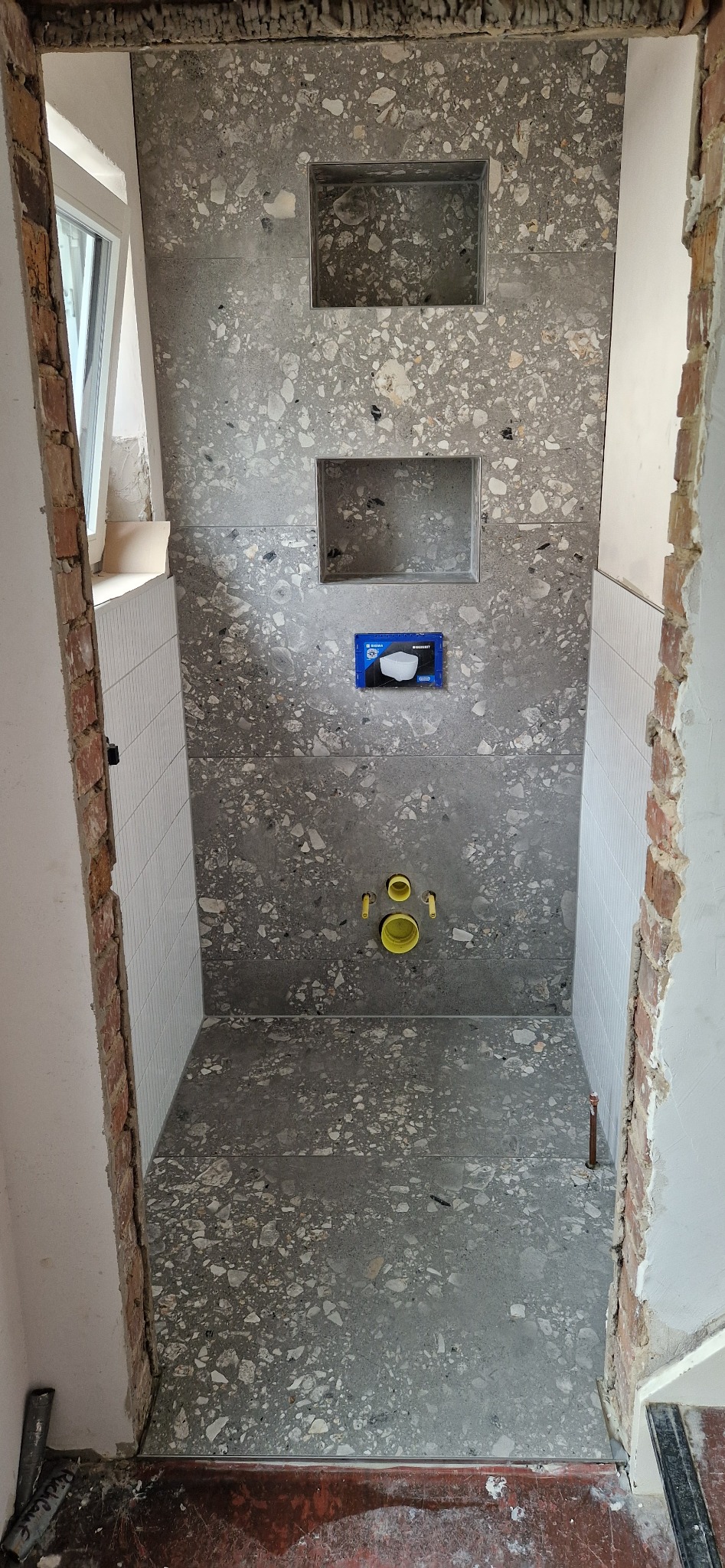 Łazienka w trakcie remontu: szare płytki lastryko na ścianach z wnękami, białe płytki na bocznych ścianach, żółte rury instalacyjne. Widoczne niedokończone wykończenia.