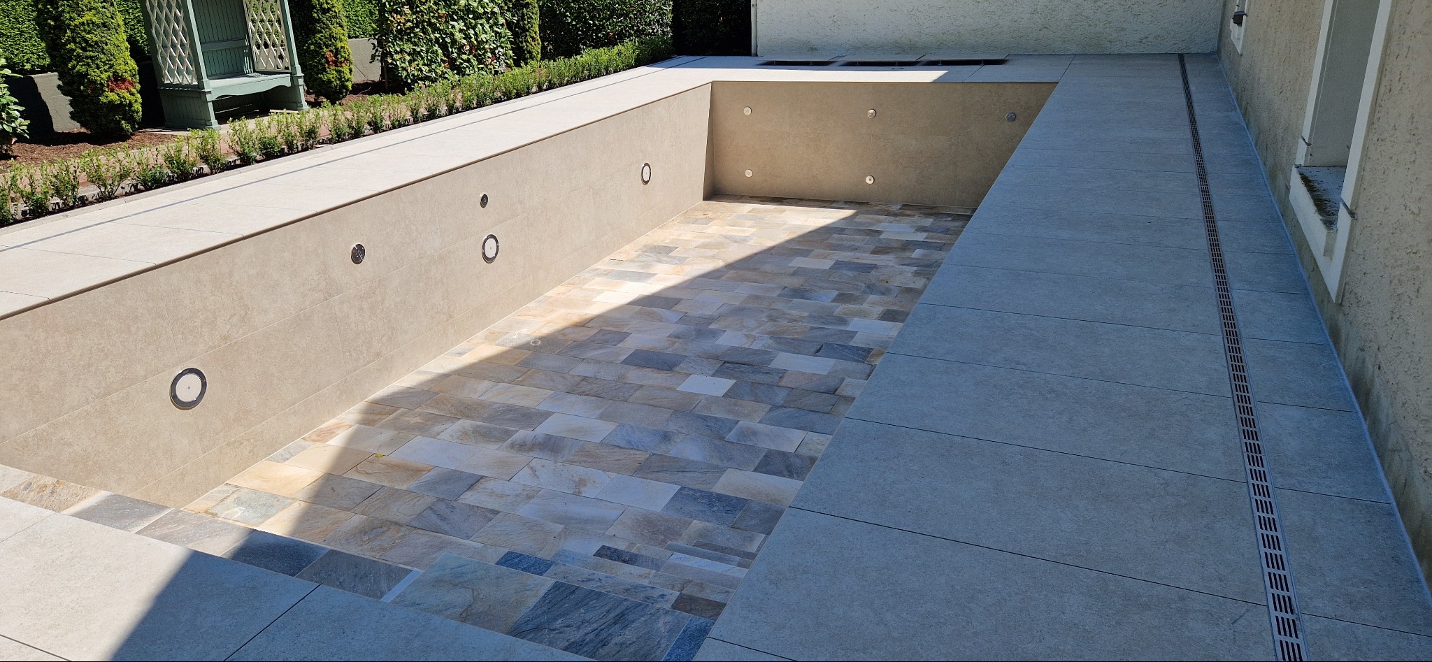 Wykończony basen z mozaiką w odcieniach beżu i szarości, oświetleniem punktowym w ścianach, otoczony jasnymi płytami i zielenią. Widoczny odpływ liniowy.