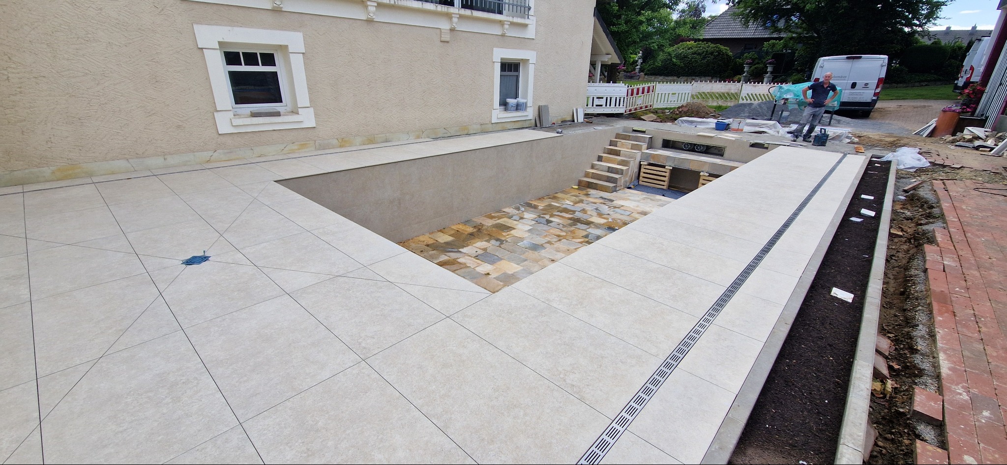 Wykończony taras z beżowych płytek z odpływem liniowym. W tle zejście do ogrodu z kamienną posadzką i schodami. Prace wykończeniowe w toku.