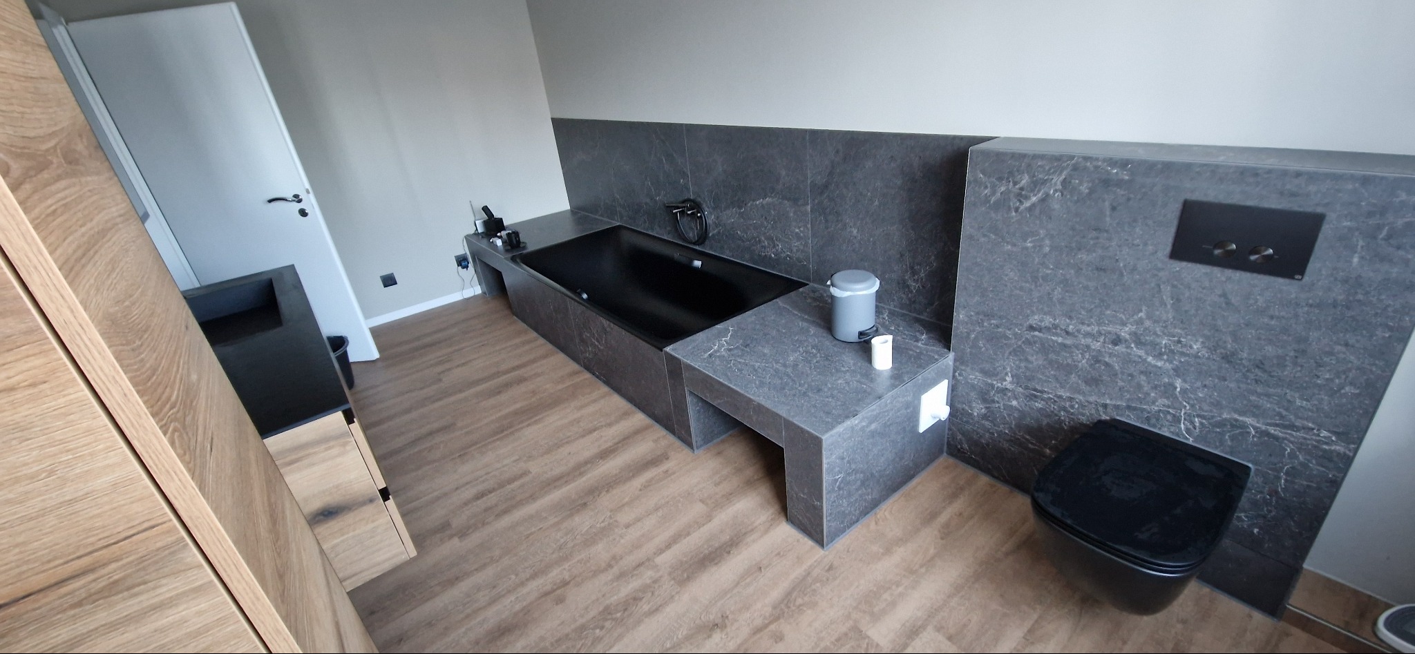 Nowoczesna łazienka z czarną wanną i toaletą, szare płytki imitujące marmur, drewniane panele podłogowe. Minimalistyczny design, neutralna kolorystyka.