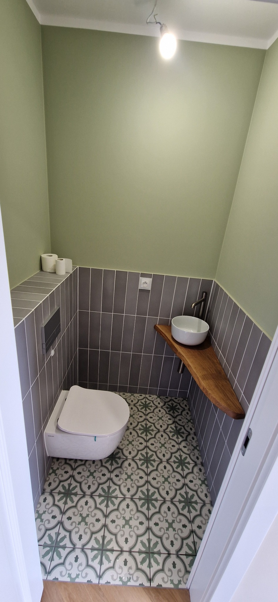 Kompaktowa łazienka z toaletą podwieszaną, umywalką na drewnianym blacie i wzorzystą podłogą. Ściany wykończone szarą glazurą i zieloną farbą.