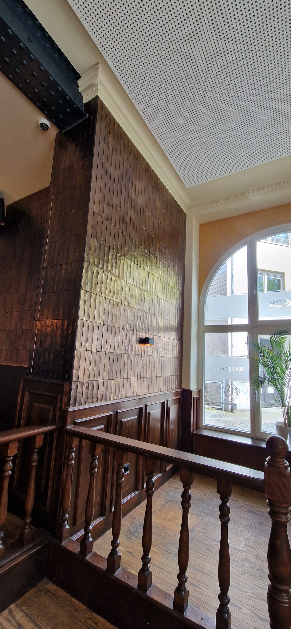 Wnętrze z brązowymi, wąskimi płytkami na ścianie, drewnianą balustradą i perforowanym sufitem. Widoczne okno i roślina doniczkowa.