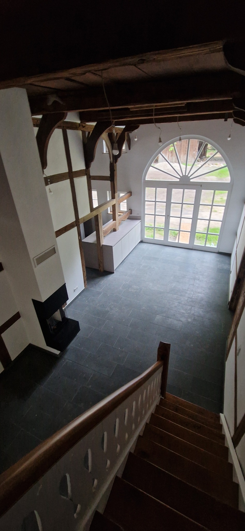 Przestronne wnętrze z szarą podłogą z płytek, drewnianą konstrukcją stropu i dużym, łukowym oknem. Widok z góry ze schodów na salon z kominkiem.