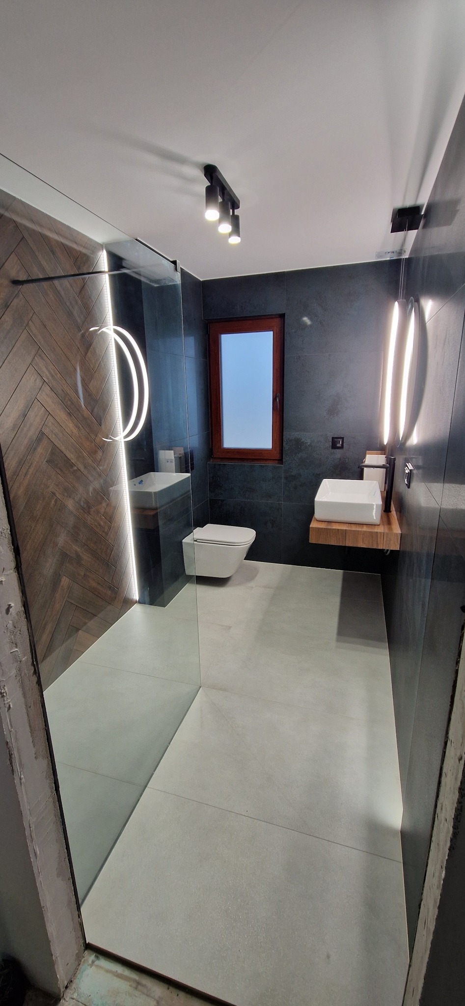 Nowoczesna łazienka z kabiną typu walk-in, drewnianą ścianą w jodełkę, podwieszaną toaletą i umywalką na drewnianym blacie. Minimalistyczne oświetlenie.