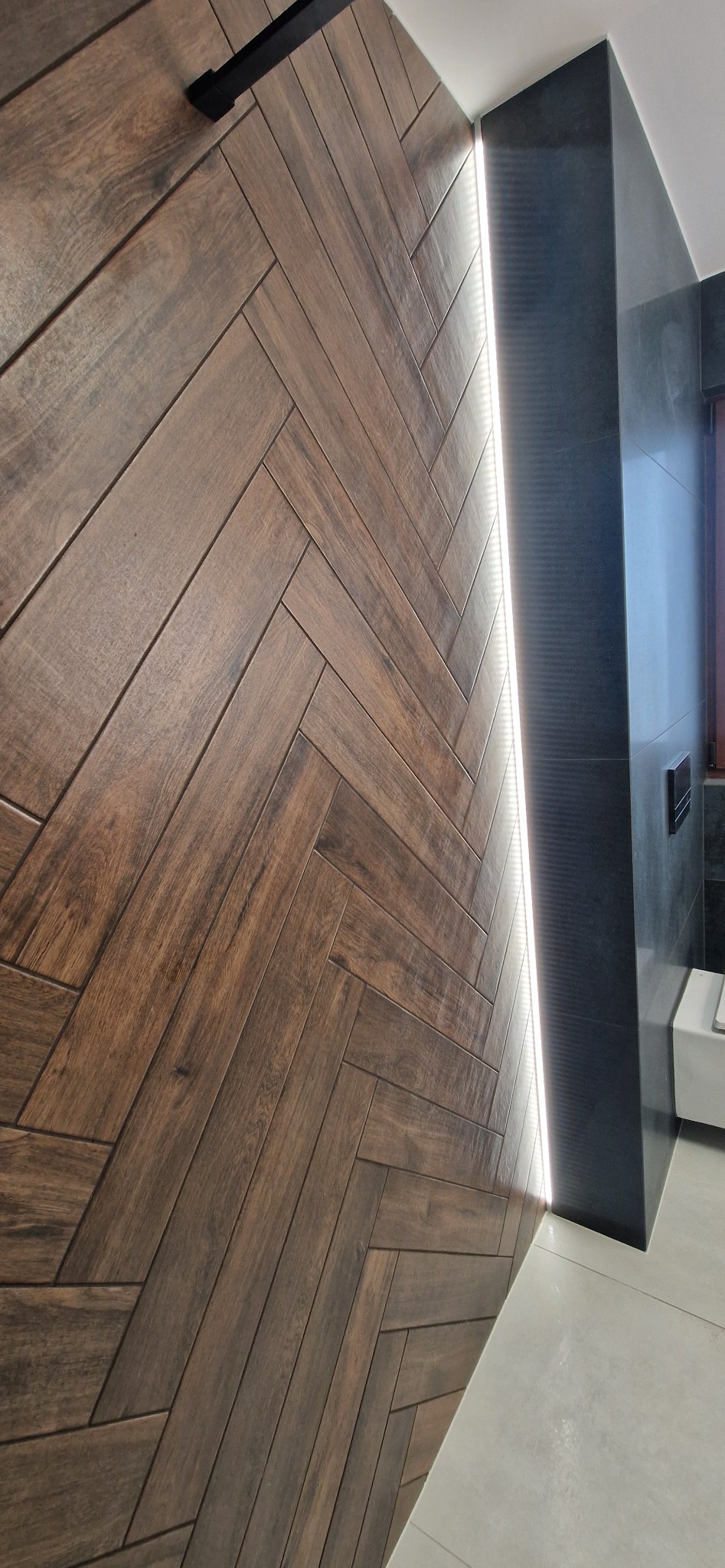 Ściana z płytek imitujących drewno ułożonych w jodełkę, podświetlona listwą LED w nowoczesnej łazience z ciemnymi elementami wykończenia.