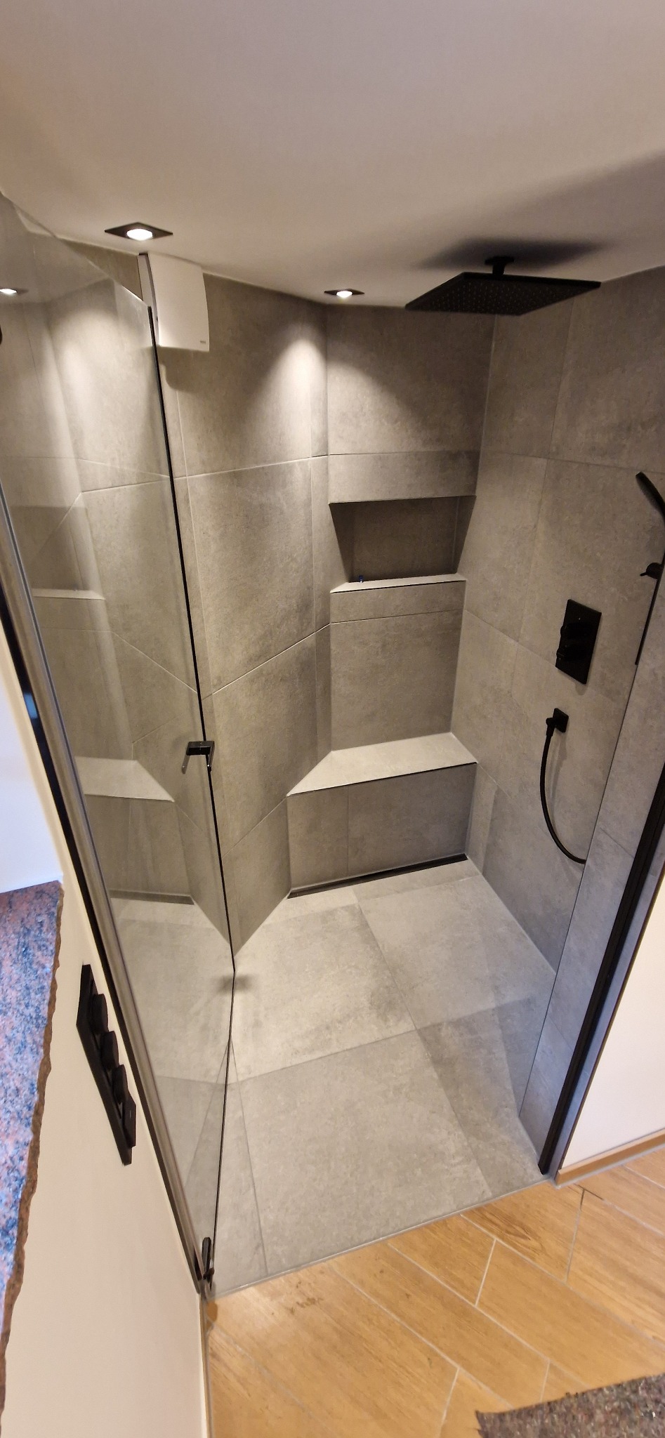 Nowoczesna kabina prysznicowa walk-in z szarymi płytkami, czarną armaturą i wnękami na kosmetyki. Minimalistyczny design, oświetlenie punktowe.