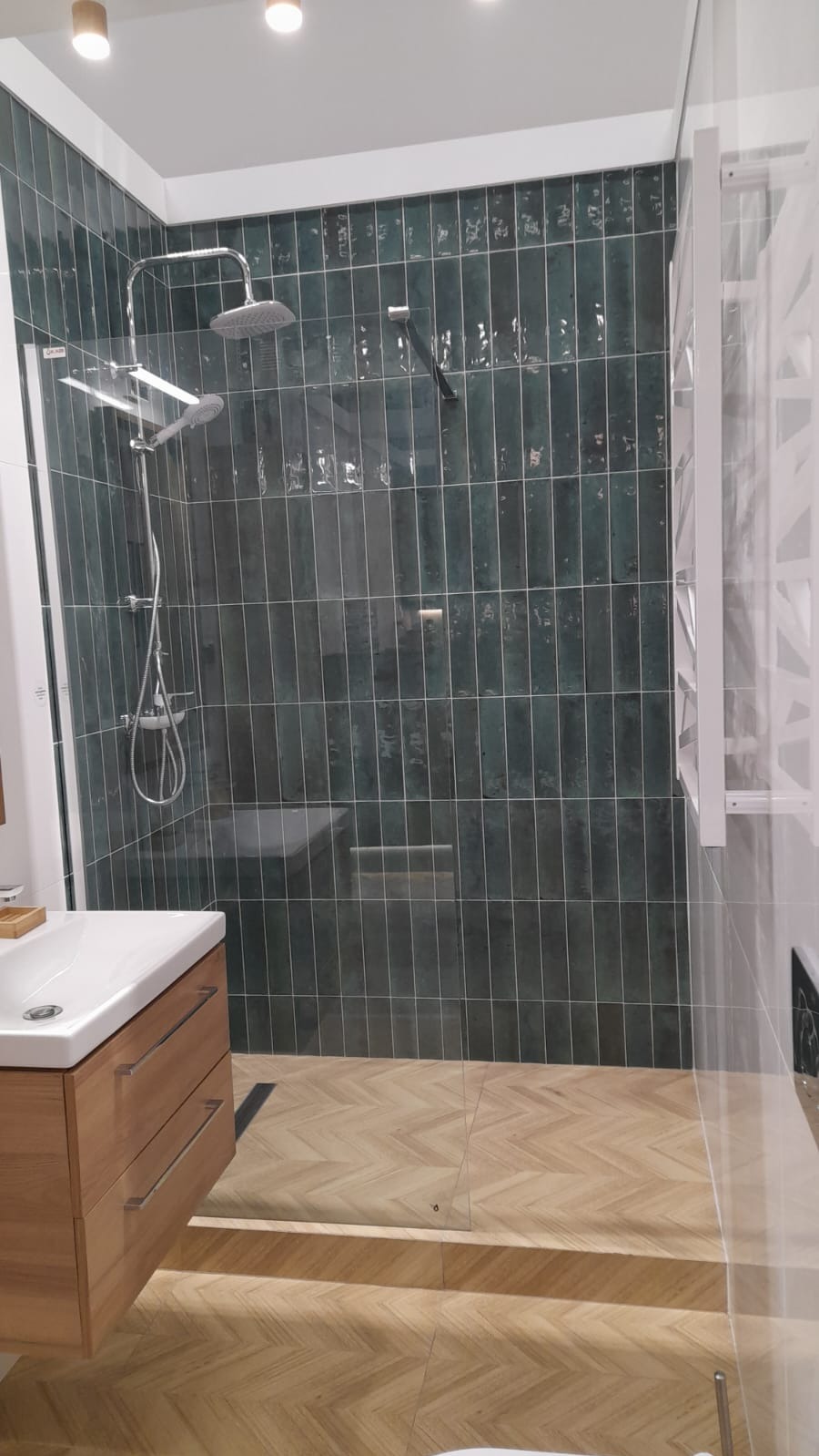 Nowoczesna łazienka z kabiną prysznicową, zielonymi płytkami i drewnianą podłogą ułożoną w jodełkę. Widoczna umywalka z szafką.