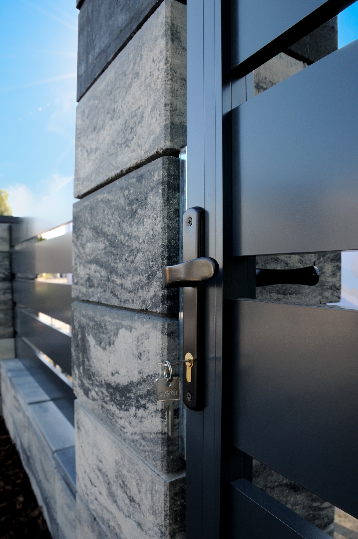 Nowoczesna brama wjazdowa z metalowymi panelami i słupkami z szarego kamienia, klucz w zamku.