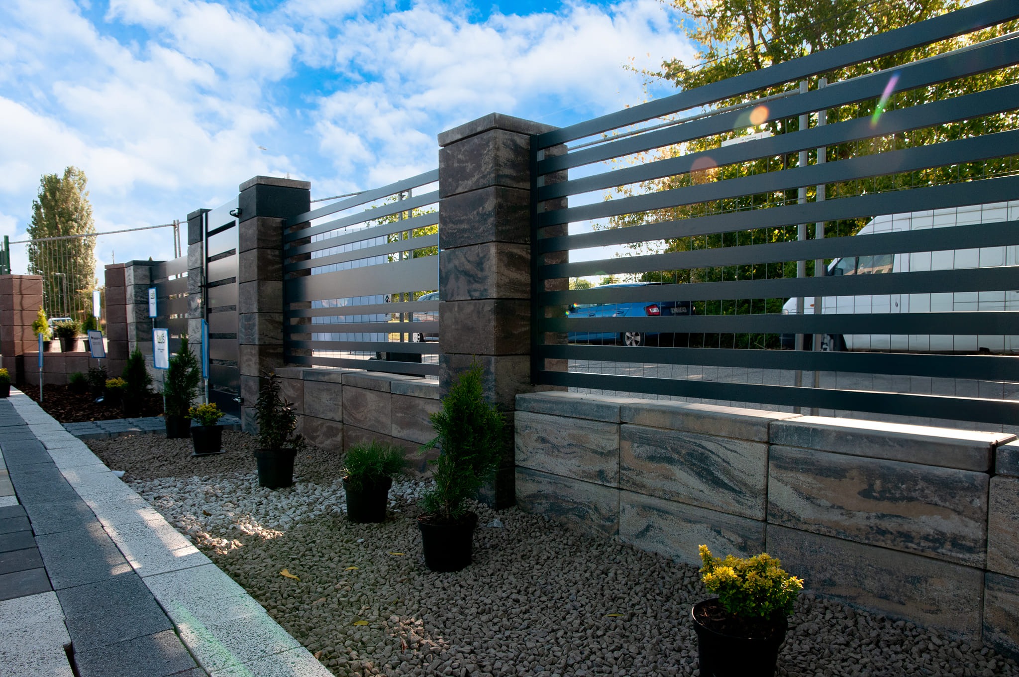 Nowoczesne ogrodzenie z poziomych metalowych profili, wsparte na słupkach z kamiennych bloków o wyraźnym wzorze, z roślinami w donicach wzdłuż ścieżki z kostki brukowej i żwirowego podłoża.