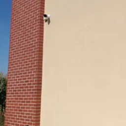 Kamera monitoringu zamontowana na elewacji budynku z ceglanym narożnikiem, obok stoi drewniana ławka.