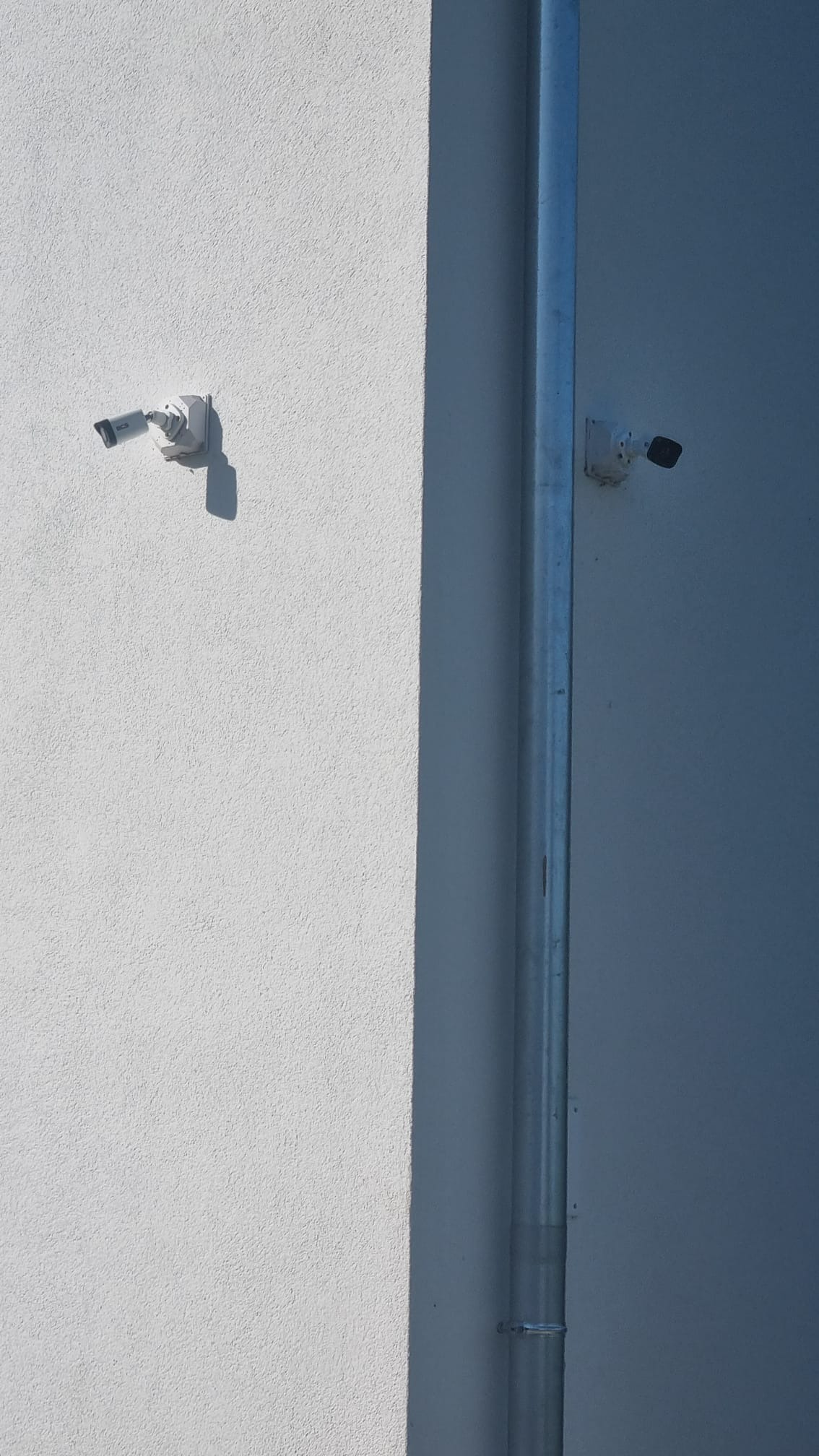 Dwie kamery monitoringu zamontowane na białej, chropowatej ścianie zewnętrznej budynku, oddzielone pionową rurą spustową. Jedna kamera po lewej, druga po prawej stronie rury.