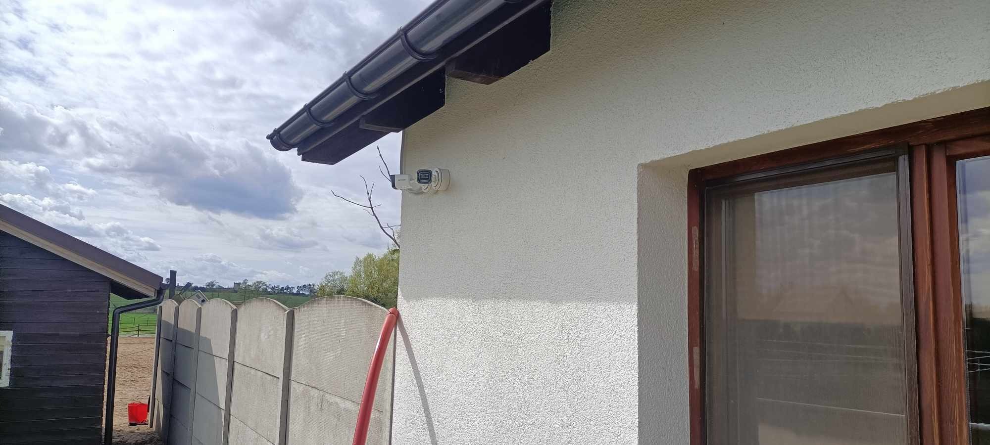 Zewnętrzna kamera monitoringu zamontowana na białej elewacji budynku z brązowymi oknami, widoczny fragment dachu z rynną, w tle ogrodzenie i zieleń