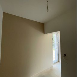 Patryk Zieliński - Pokój po częściowym pomalowaniu ścian na dwa odcienie beżu, z widocznym kablem zwisającym z sufitu i zabezpieczonym oknem.