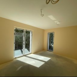 Patryk Zieliński - Pomalowane na beżowo wnętrze pokoju z dużymi oknami balkonowymi i jednym mniejszym oknem, widoczne kable wystające z sufitu, podłoga zabezpieczona folią malarską.