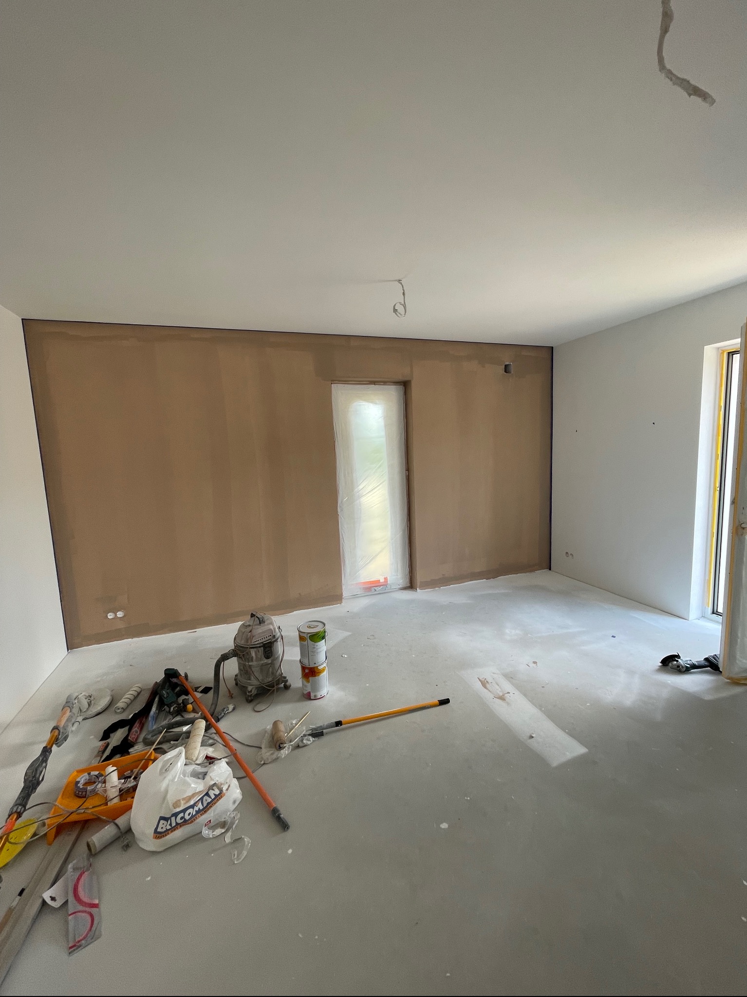 Pokój w trakcie remontu, ściana częściowo pokryta gładzią, na podłodze narzędzia i materiały budowlane, w tle drzwi zabezpieczone folią.
