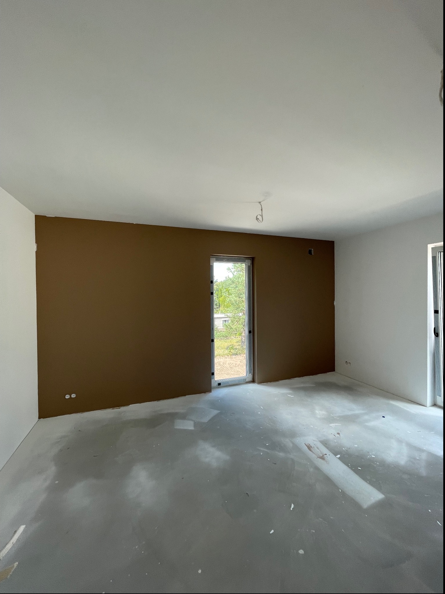 Pokój w trakcie remontu z jedną ścianą pomalowaną na brązowo, surowy beton na podłodze, widoczne niedoskonałości i ślady po pracach, okno balkonowe z widokiem na zieleń.