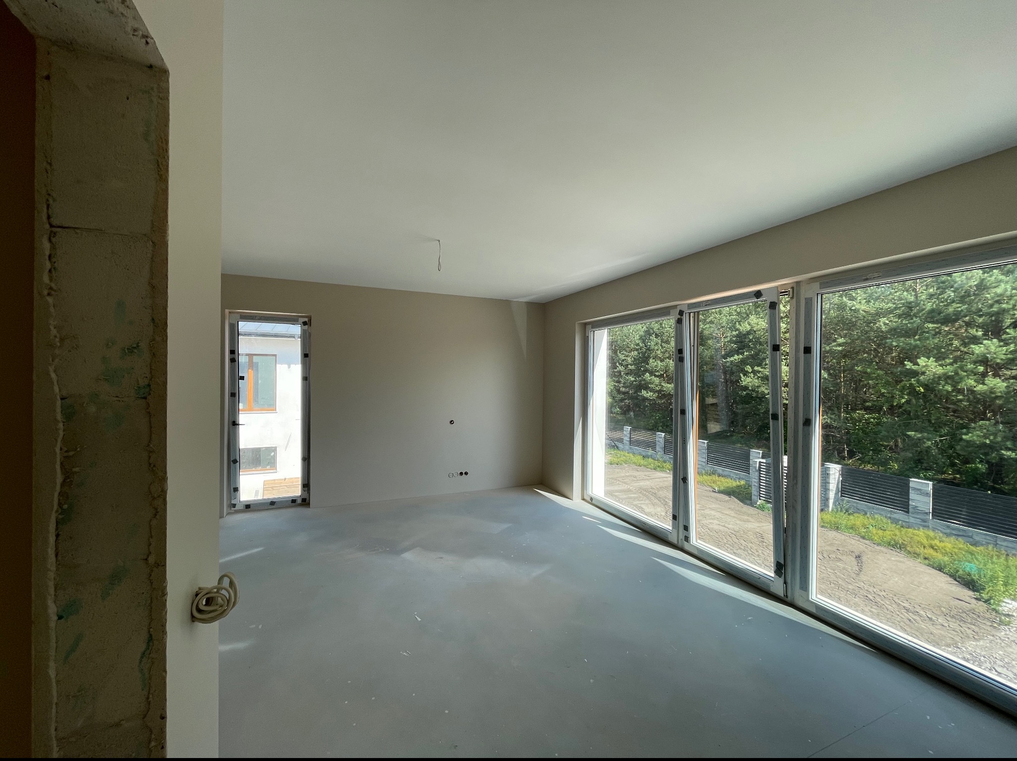Surowe wnętrze pokoju z dużymi oknami, widoczne ślady po szpachlowaniu na podłodze i ścianach, wystające kable elektryczne, widok na las za oknem.