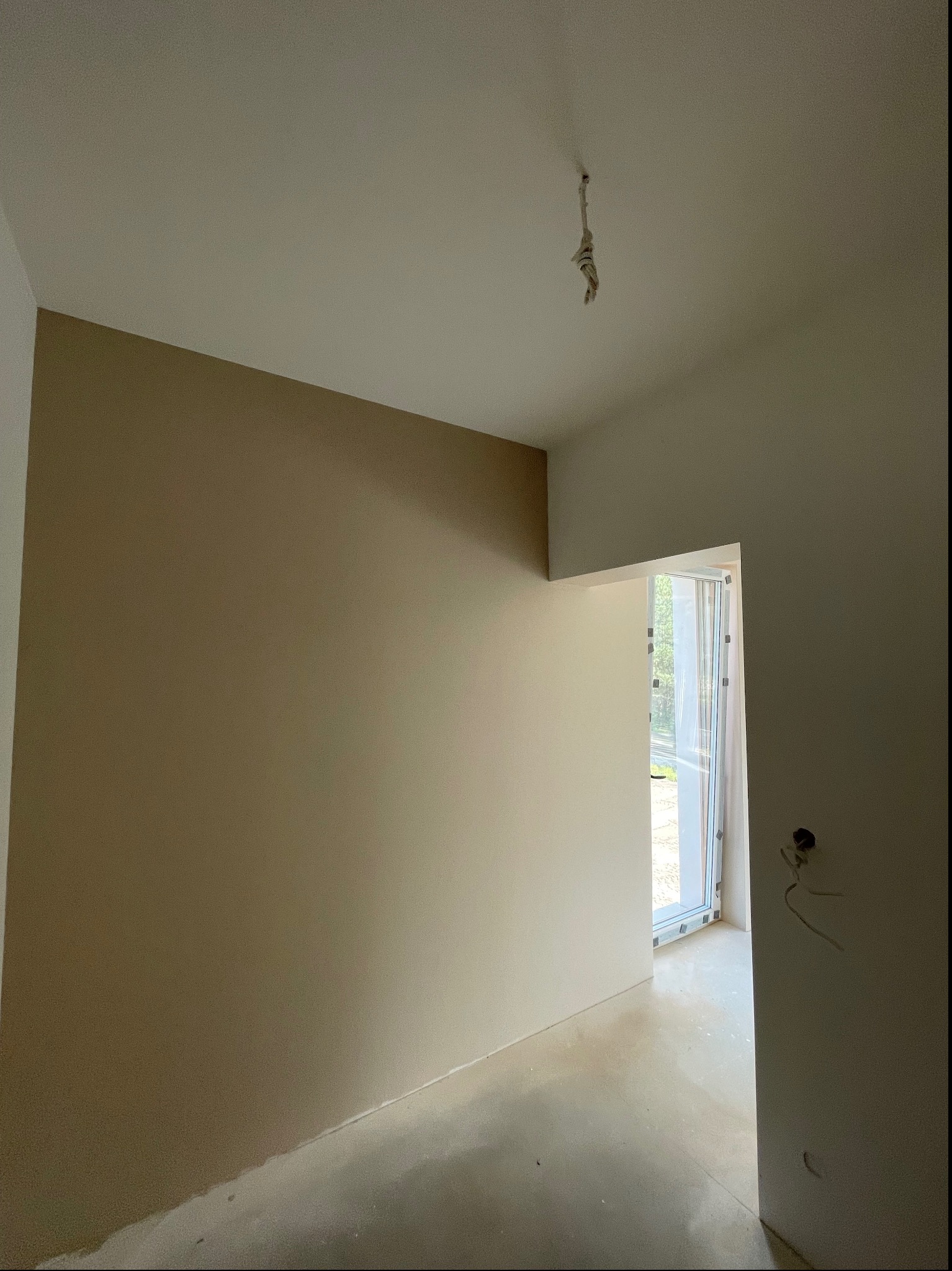 Pokój po częściowym pomalowaniu ścian na dwa odcienie beżu, z widocznym kablem zwisającym z sufitu i zabezpieczonym oknem.