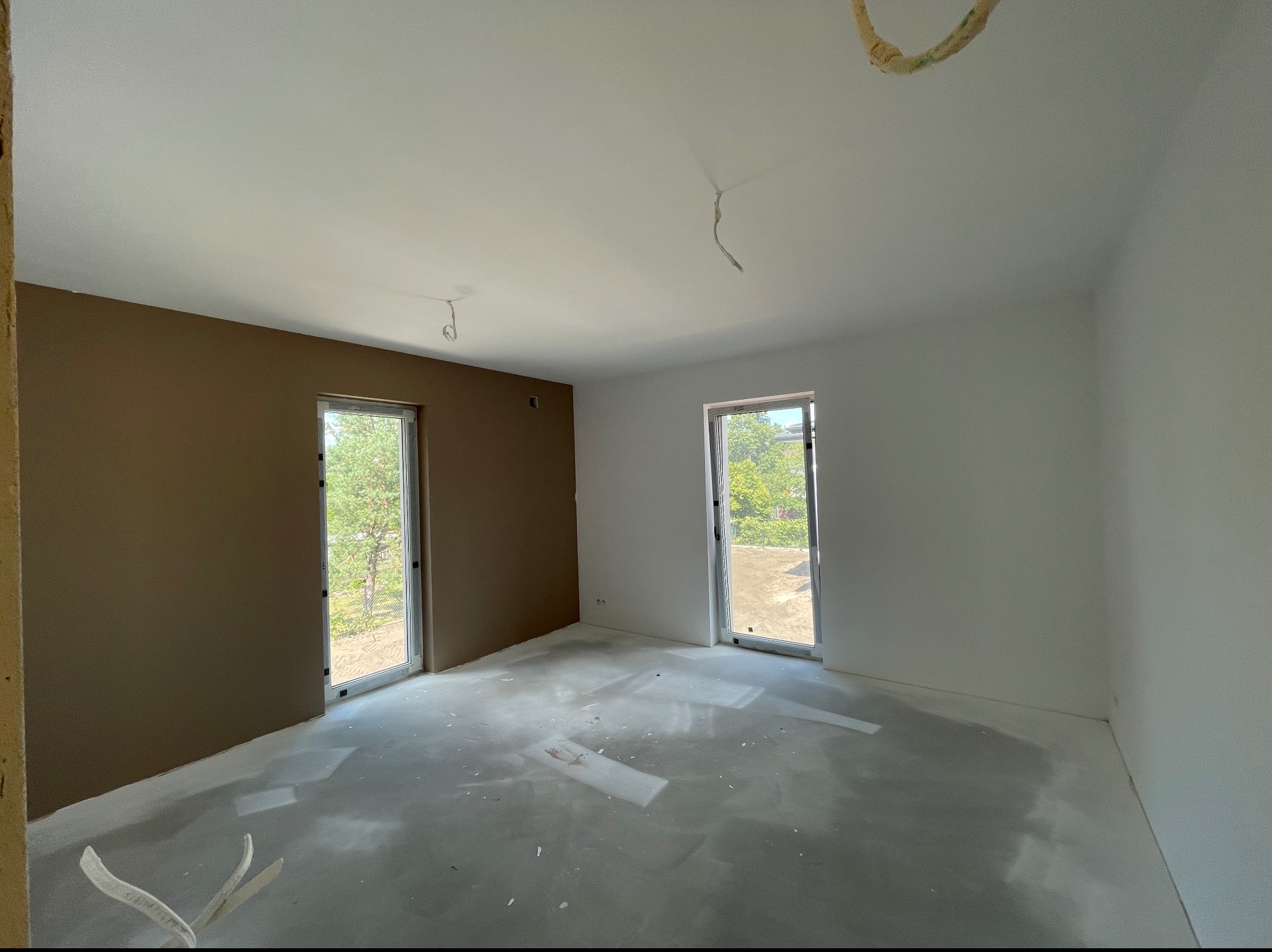 Pokój w trakcie prac wykończeniowych: jedna ściana pomalowana na brązowo, pozostałe na biało, widoczne ślady po szpachlowaniu na podłodze, okna z widokiem na zieleń.